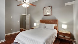 Un dormitorio con una cama grande, cabecera de madera, dos mesitas de noche con lámparas, un ventilador de techo y una puerta que da a otra habitación.