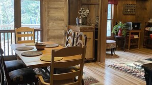 Uma área de jantar de madeira com mesa e cadeiras, uma cozinha com geladeira e um laptop, e vista para o exterior através de uma porta.