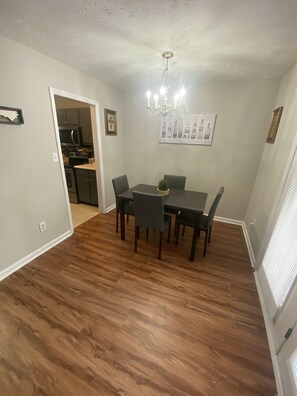 A dining area with a table and chairs, a chandelier, and a kitchen in the background.