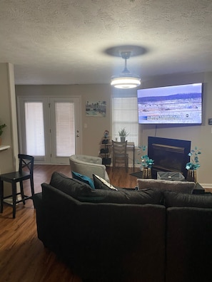 A living room with a dark-colored sofa, a dining table with chairs, a television mounted on the wall, and a ceiling light fixture.