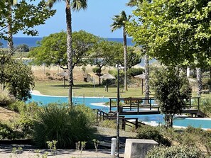 A pool area with a wooden deck, surrounded by lush greenery and palm trees, overlooking a body of water.