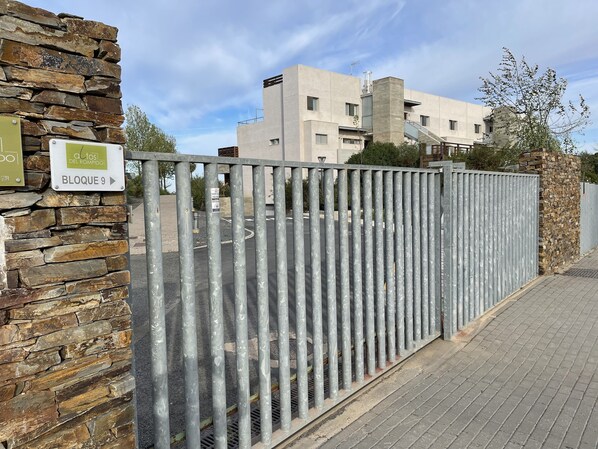 A gated entrance with a stone wall and a metal fence, leading to a modern building with multiple windows and balconies.