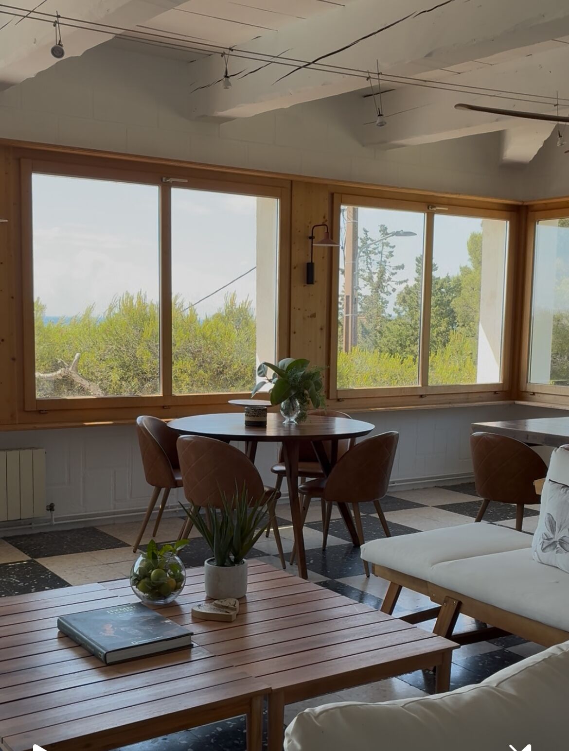 A dining area with a round table and chairs, a sofa, and a view of greenery through large windows.