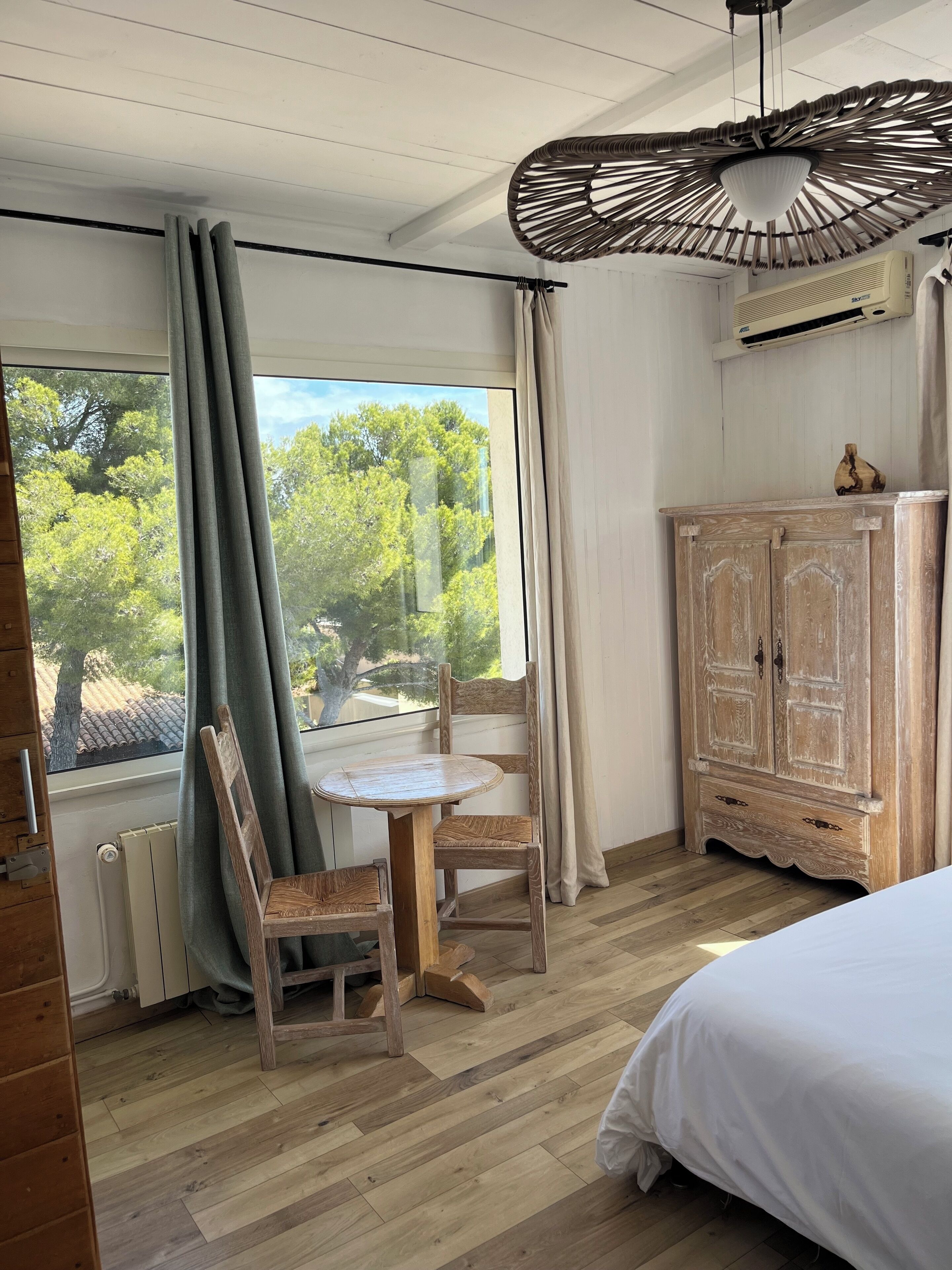 A room with a wooden floor, a wooden chair, a small round table, a bed, a wardrobe, and a window with curtains.