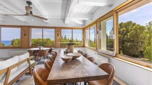 Ein geräumiger Essbereich mit einem langen Holztisch und Ledersesseln, große Fenster mit Blick auf das Meer und viel Grün, sowie ein Deckenventilator.