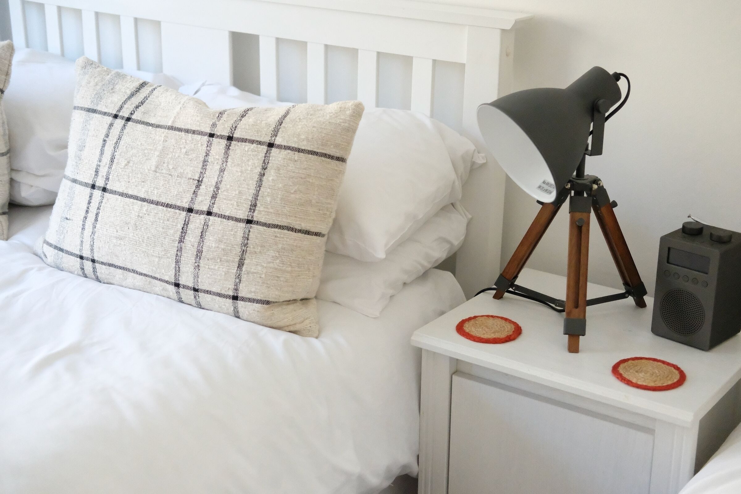 Un lit bien fait, avec une literie blanche et un coussin à carreaux, une lampe de chevet posée sur une table de nuit blanche, et une petite enceinte.