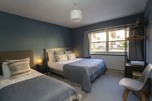 A bedroom with two beds, a bedside table with a lamp, a chair, a window with curtains, and a radiator.