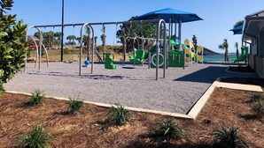 A playground with swings, slides, and climbing structures, surrounded by a sandy area and small plants. - Emerald Coast Stay! (Navarre)