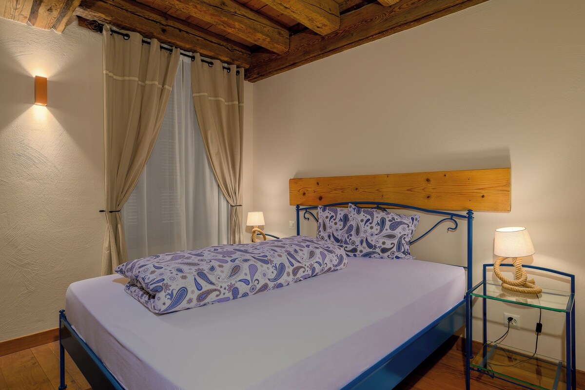 Un dormitorio con cabecera de madera, cama con ropa de cama estampada, dos lámparas de noche y una mesita de noche de cristal con una lámpara.