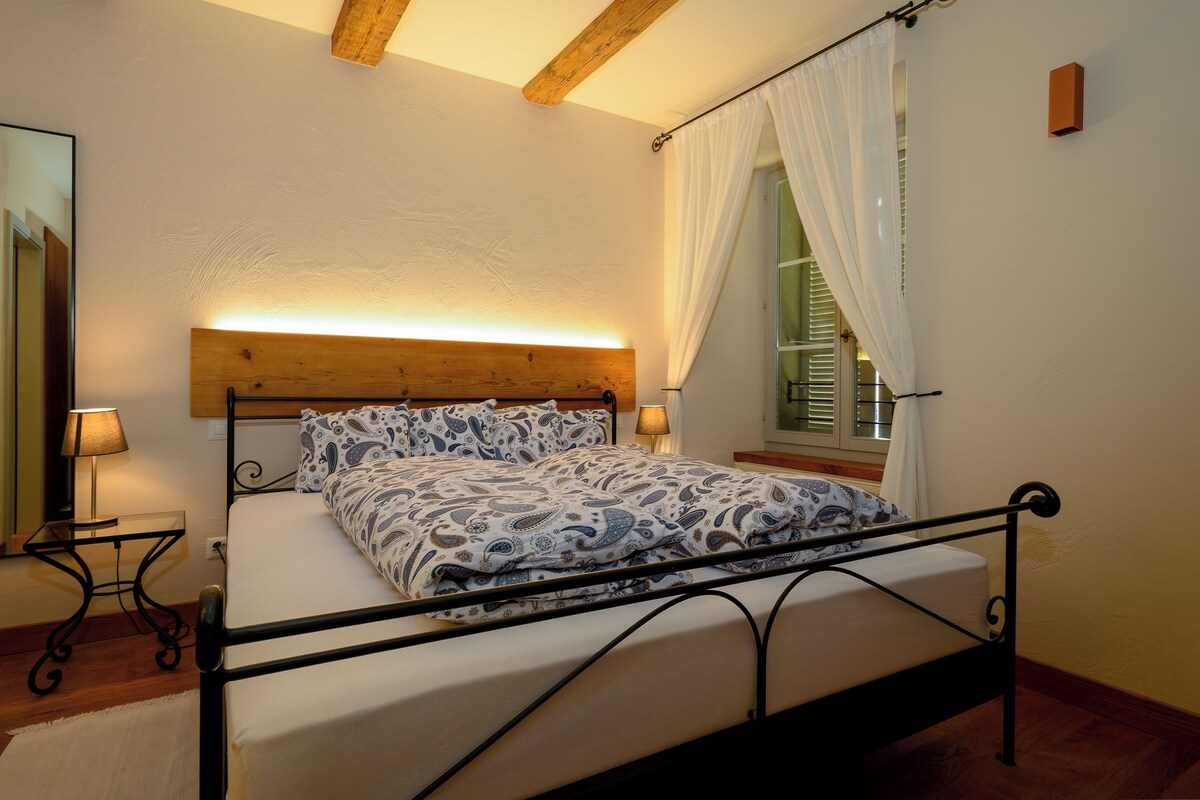 Un dormitorio con cama, lámparas de noche, cabecera de madera y una ventana con cortinas.