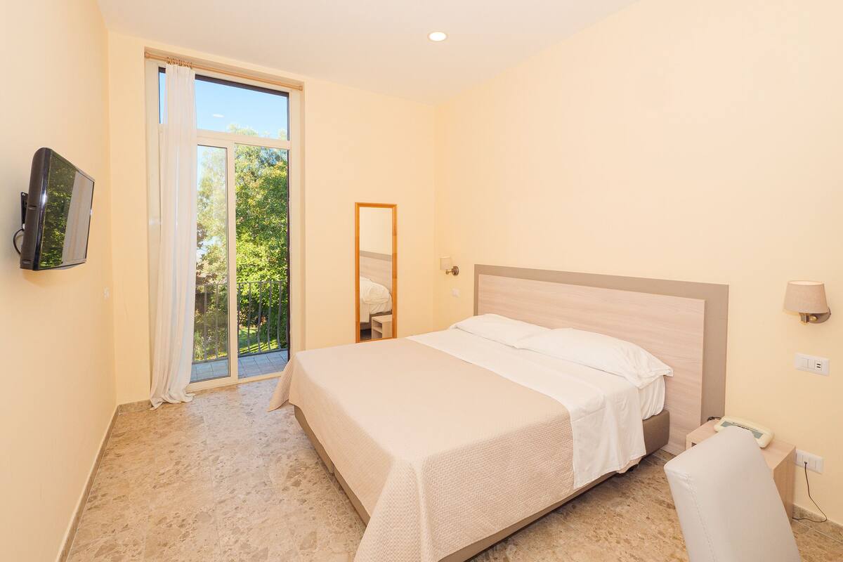 Une chambre à coucher avec un lit, un téléviseur fixé au mur, une lampe de chevet et une fenêtre donnant sur un paysage verdoyant.