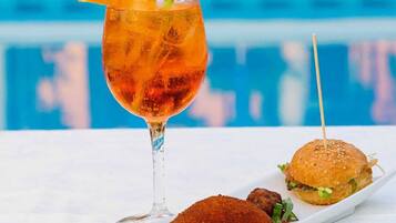 Um copo de chá gelado com uma fatia de laranja e um canudo, um hambúrguer com pão de gergelim e uma coxinha de frango frita em um prato branco, tudo sobre uma mesa perto de uma piscina.