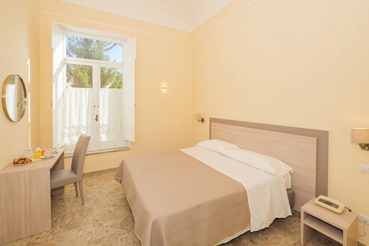 Une chambre d’hôtel comprenant un lit, un bureau avec des articles pour le petit-déjeuner, une chaise, une table de chevet avec une lampe et une fenêtre avec des rideaux.