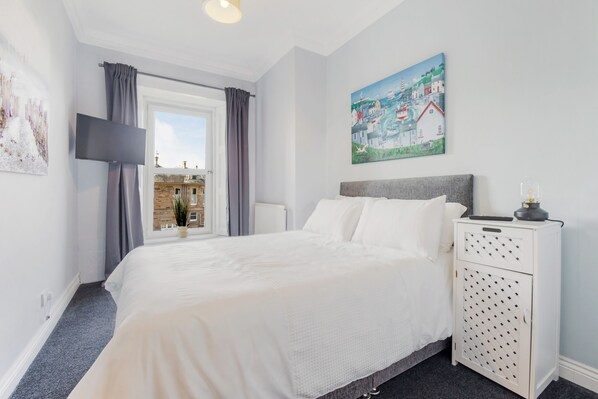 A bedroom with a large bed, a nightstand with drawers, a flat-screen TV, and a window with curtains.