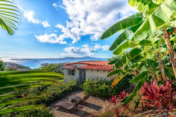 A house with a red-tiled roof, a patio with a table and chairs, and a view of the sea and mountains.