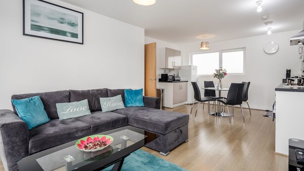 A modern living room with a grey sectional sofa, a glass coffee table, and a dining area with a table and chairs. - Impeccable 2-bed Apartment in Romford (Romford)