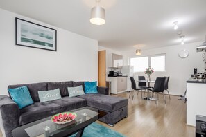 A modern living room with a grey sectional sofa, a glass coffee table, and a dining area with a table and chairs. - Impeccable 2-bed Apartment in Romford (Romford)