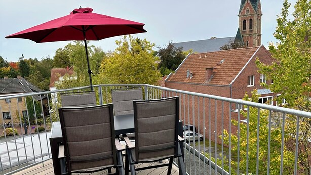 A wooden deck with a table and chairs, a red umbrella, and a view of a church steeple in the background.