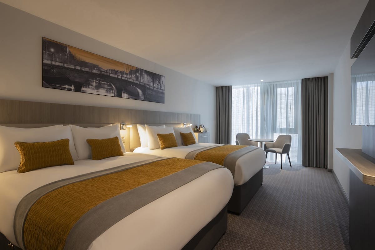 A hotel room with two beds, a desk, a chair, and a large window with curtains.