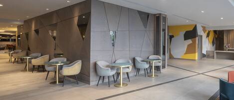 Un interior moderno con una zona de estar que incluye mesas y sillas redondas, un diseño geométrico en la pared y un techo visible con iluminación empotrada.