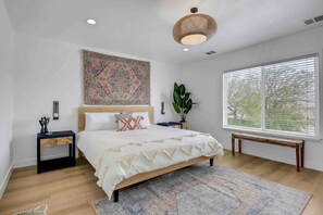 A bedroom with a large bed, a patterned tapestry, a potted plant, and a large window with blinds.