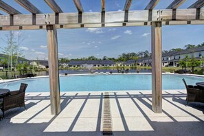 Una pérgola de madera sobre una piscina con sillones de descanso, vista despejada de la piscina y zona residencial al fondo.