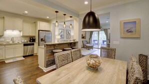 Une cuisine spacieuse avec des meubles blancs, un îlot central doté de tabourets et un coin repas avec une table en bois et des chaises en osier.