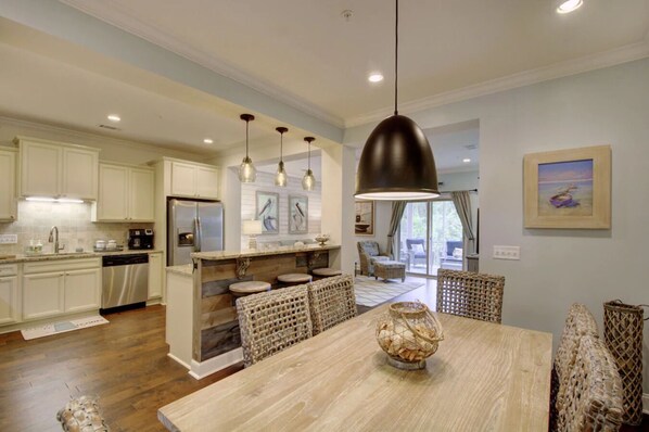 Una cocina amplia con armarios blancos, una isla central con taburetes y una zona de comedor con mesa de madera y sillas de mimbre.