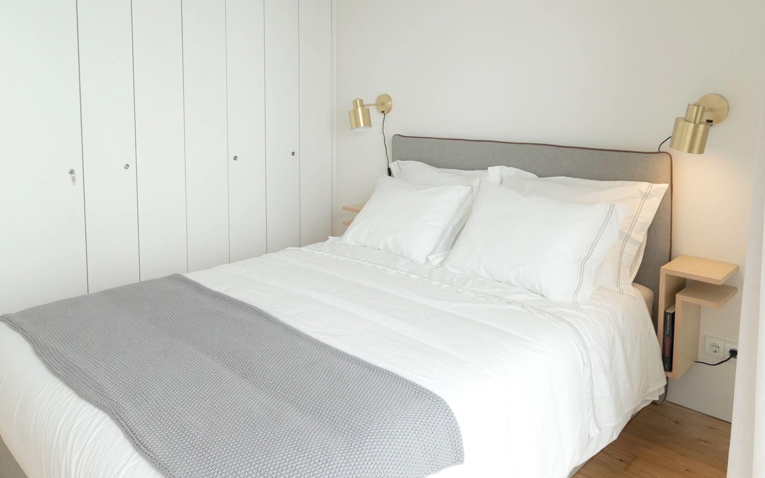 Un lit bien fait, avec du linge de lit blanc et un plaid gris, une table de chevet en bois avec une lampe, et une armoire blanche avec des poignées verticales.