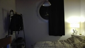 Ein schwach beleuchteter Raum mit einem Bett, einem kleinen Tisch mit Lampe, einem Fenster mit Vorhang und einem Kleiderständer mit Kleidung.