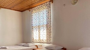 A hotel room with three single beds, each with a white bedspread and pillows, a wooden ceiling, and a window with a polka dot curtain.