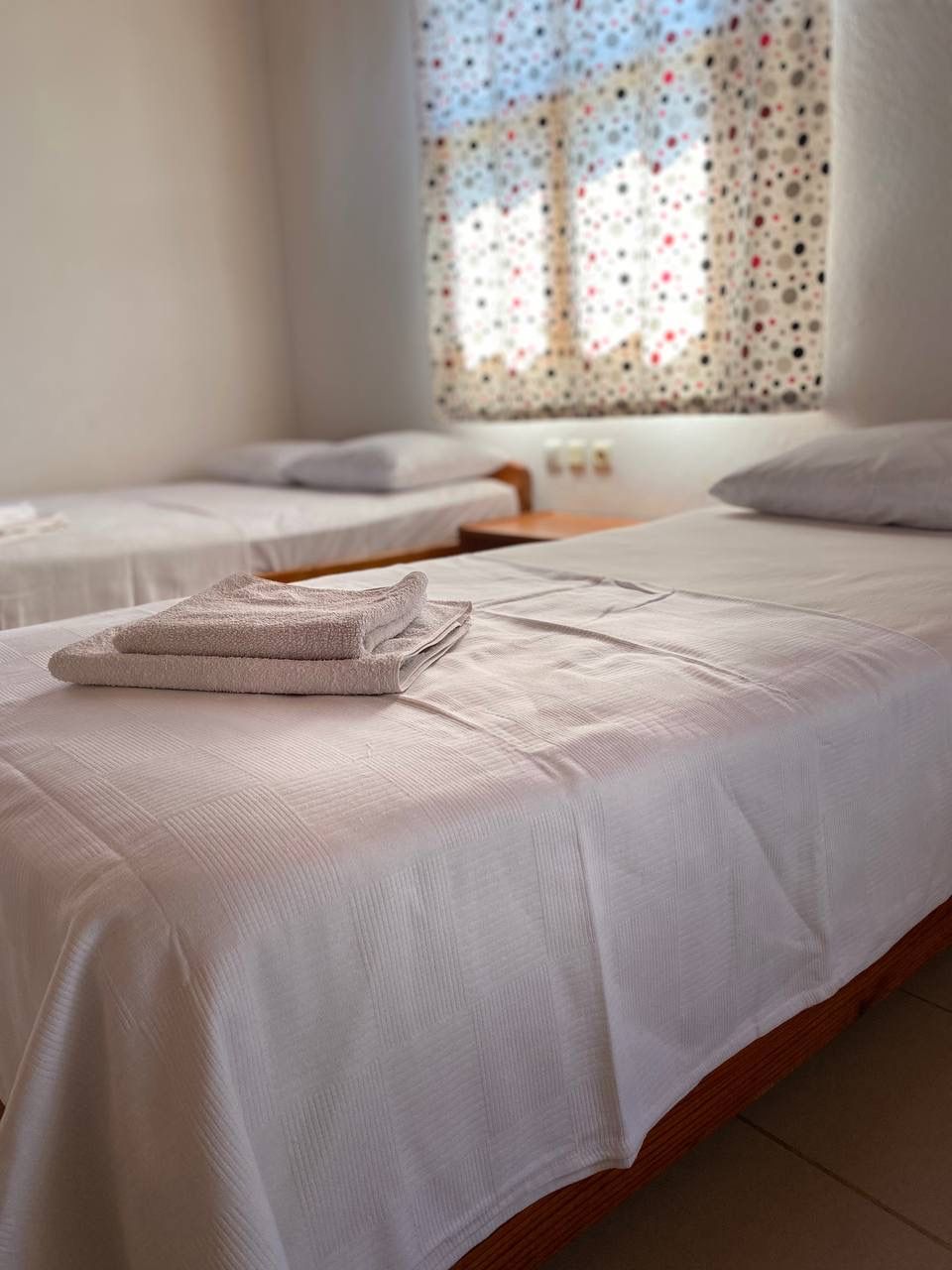 Habitación de hotel con dos camas, cada una con una toalla doblada con cuidado y una colcha blanca.