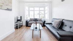 A living room with a glass coffee table, a black sofa, a grey sofa, a TV on a stand, and a large window with a view of houses. - Charming 3-bed Apartment in Romford (Romford)