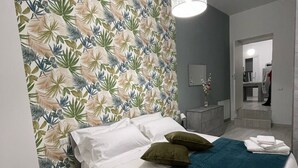 Een moderne hotelkamer met een groot bed, een smaragdgroene gewatteerde spreideken, witte lakens en behangpapier met een tropisch bladmotief.