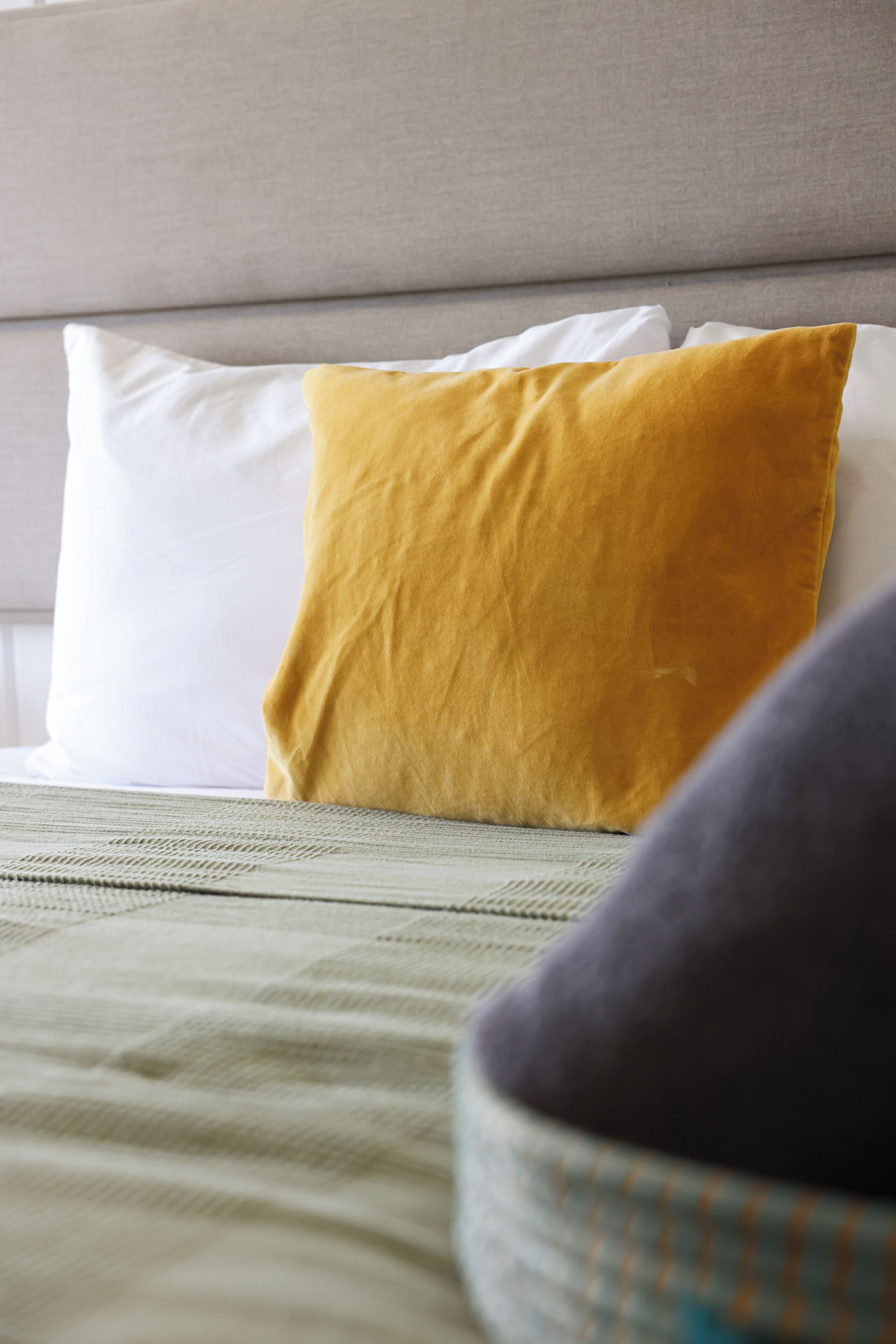 Una cama con un almohadón blanco y uno mostaza, una manta verde y un cabecero gris.