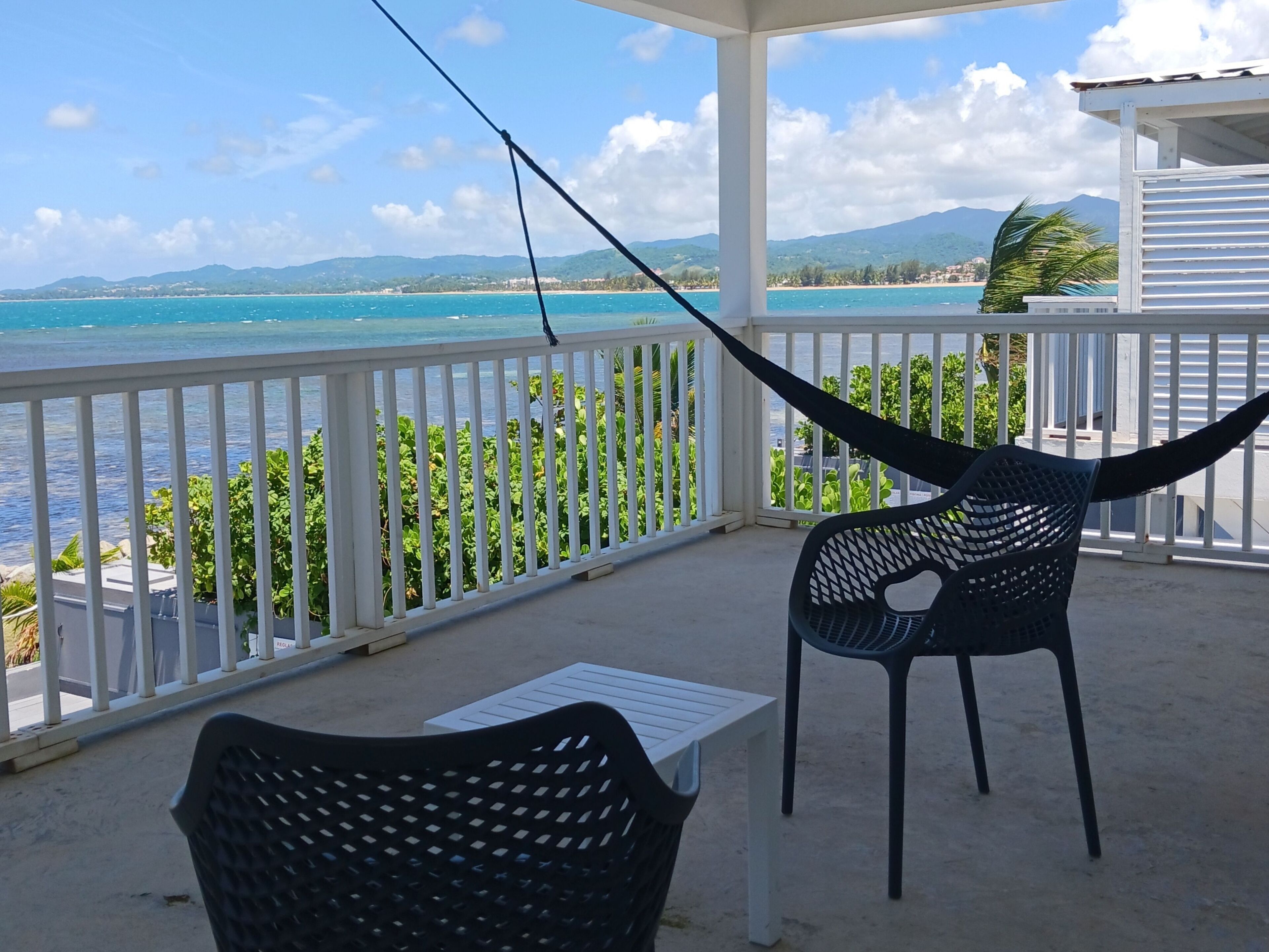 A balcony with a view of the ocean and mountains, a hammock, and a table.