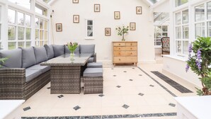 A conservatory with a sectional sofa, a wooden chest of drawers, and framed pictures on the walls. - The Abbey Cottage (Bury St Edmunds)