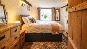 A bedroom with a wooden bed, a dresser, a mirror, and a window with curtains. - The Abbey Cottage (Bury St Edmunds)