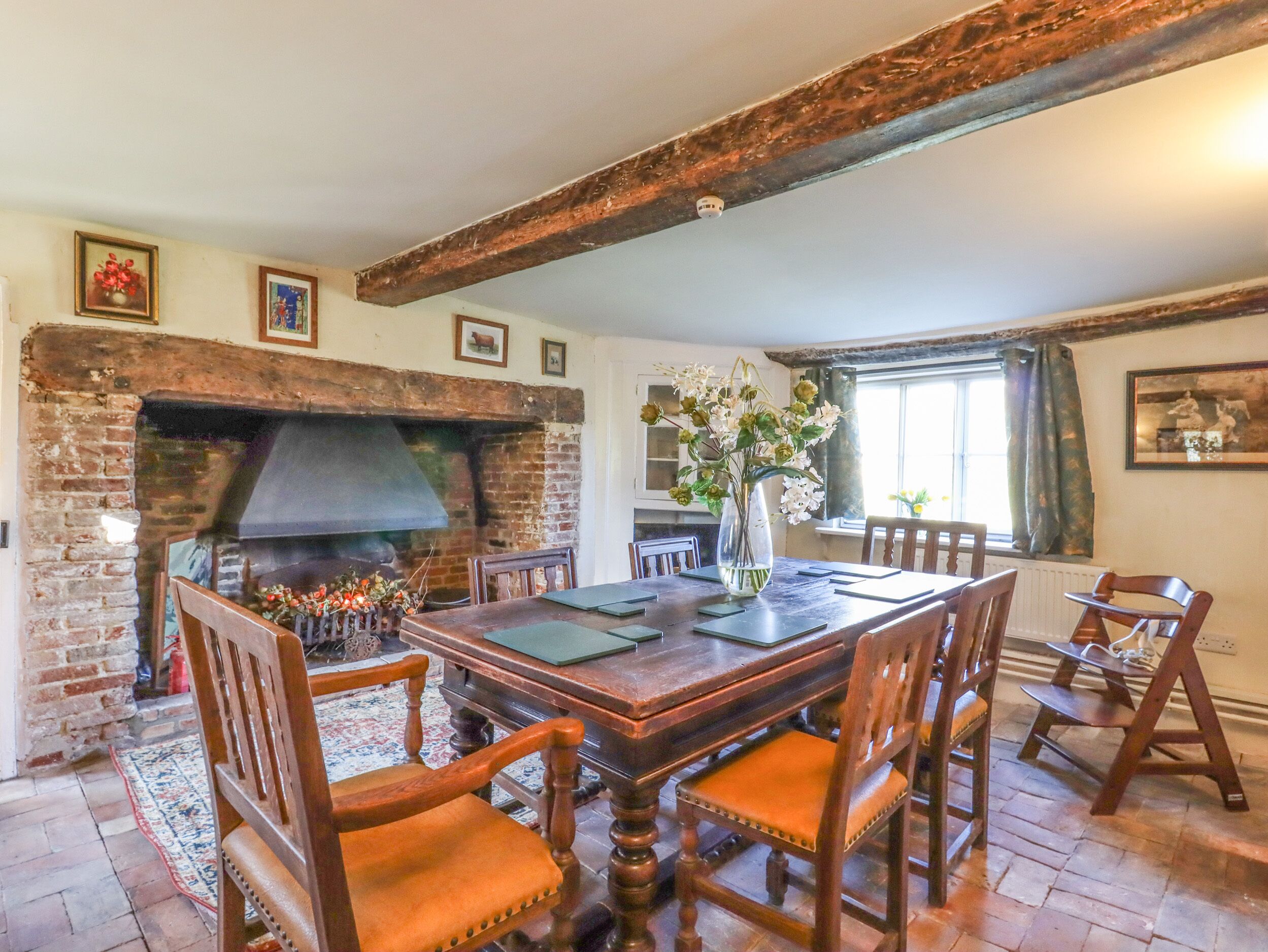 A dining room with a brick fireplace, wooden chairs, a table, and a vase with flowers.
