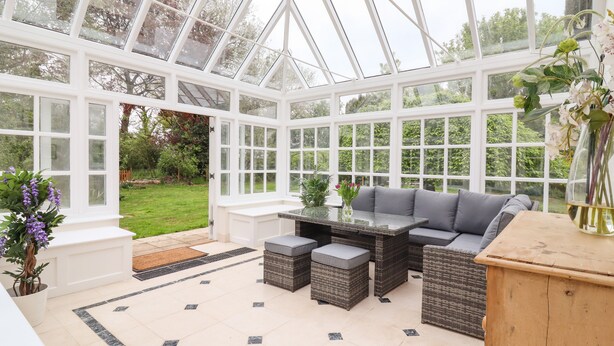 A conservatory with a glass ceiling, a seating area with cushions, and a view of the garden. - The Abbey Cottage (Bury St Edmunds)