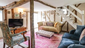 A rustic living room with exposed wooden beams, a television, a patterned armchair, a tufted ottoman, and a blue sofa. - The Abbey Cottage (Bury St Edmunds)
