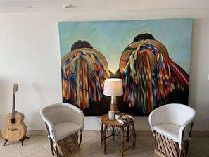 A room with two white armchairs, a small round table with a lamp, and a guitar against a wall with a colorful painting.