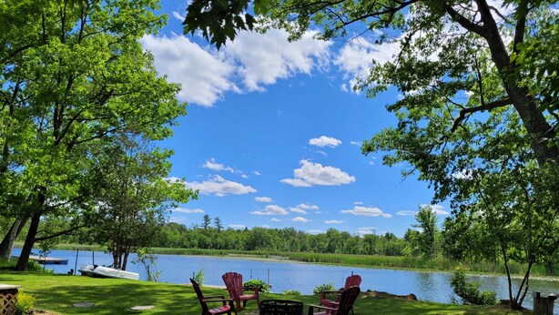 Eine Terrasse am Seeufer mit Adirondack-Stühlen und einem Tisch, umgeben von üppigem Grün und blauem Himmel.