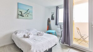 Una cama bien hecha con sábanas blancas, una silla azul, una mesita y un cuadro de pájaros en la pared.