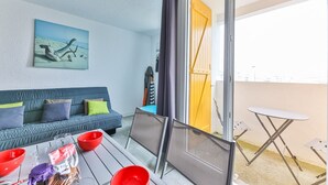 Una habitación con un sofá azul, una mesa con cuencos rojos, un cuadro de una playa y un ventanal corredizo que da a un espacio exterior.