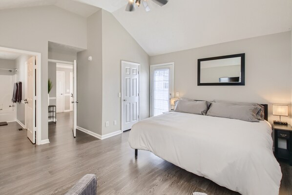 A bedroom with a large bed, a ceiling fan, a mirror, and a door leading to another room.