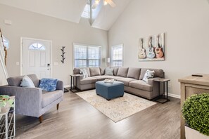 A spacious living room with a grey sofa, a blue ottoman, and a wooden coffee table. There are two windows with blinds, a white door, and a ceiling fan.