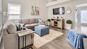 A modern living room with a sectional sofa, a blue ottoman, a flat-screen TV, and a decorative wall clock.