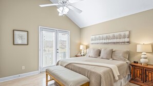 Un dormitorio con una cama grande, un banco, un ventilador de techo, un cuadro y una mesita de noche con lámparas.