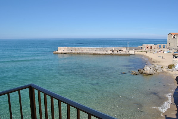 Vista costera con cielo despejado, una playa arenosa y un muelle de piedra que se adentra en el mar.